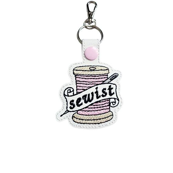 White faux leather Sewist keyring embroidered with a pink thread spool and needle design, finished with a metal clasp.