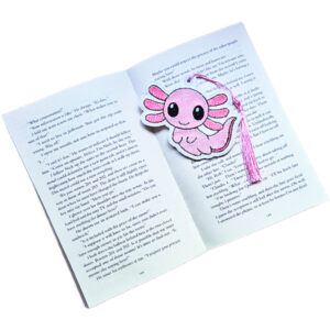 Pink embroidered axolotl bookmark with lilac tassel placed inside an open book, showing how it keeps a reader's page.