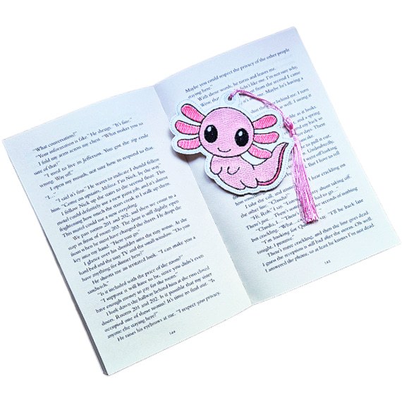 Pink embroidered axolotl bookmark with lilac tassel placed inside an open book, showing how it keeps a reader's page.