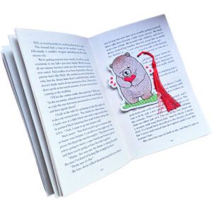 Embroidered bear bookmark holding a heart, placed inside an open book with a tassel draped over the pages to show it in use.