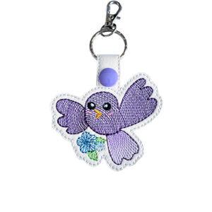 Embroidered purple kawaii bird keyring with flower detail, white faux leather strap, purple snap fastener and silver keyring clip.