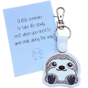 White faux leather cute sloth keyring with embroidered sloth face, shown beside a white keepsake card with gentle reminder wording.