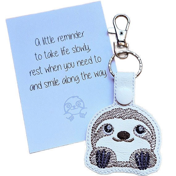White faux leather cute sloth keyring with embroidered sloth face, shown beside a white keepsake card with gentle reminder wording.
