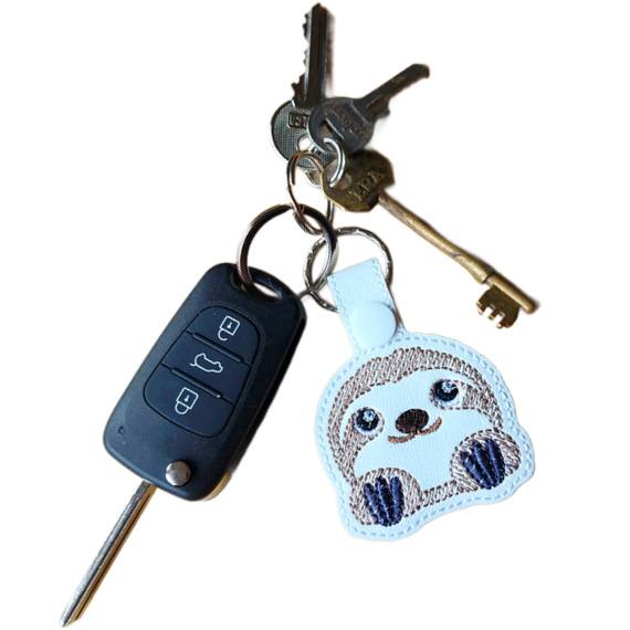 White faux leather cute sloth keyring with embroidered kawaii sloth face, shown attached to a set of keys with silver keyring hardware.