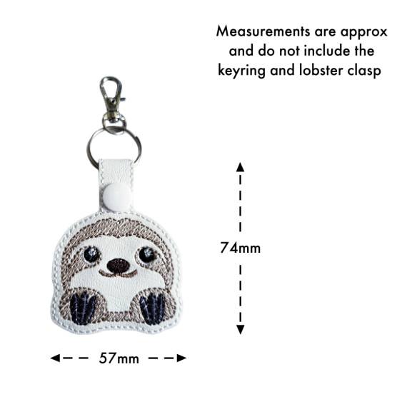 White faux leather cute sloth keyring with embroidered kawaii sloth face, shown with measurement arrows indicating an approximate size of 74mm x 57mm, excluding the keyring and lobster clasp.