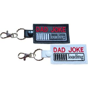 Dad Joke Loading keyrings shown in black and white faux leather, with embroidered Dad Joke lettering, loading bar design and sturdy metal keyring hardware.