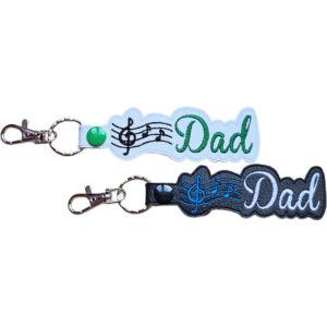 Dad Music Note keyrings shown in white and black faux leather, with embroidered musical notes, Dad lettering and sturdy metal keyring hardware.