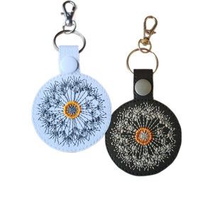 Two embroidered dandelion clock keyrings in white and black faux leather, with silver and gold clips and matching snap fasteners.