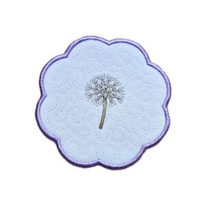 Embroidered dandelion seed head candle coaster with a single stitched dandelion clock in the centre, soft textured background and purple scalloped border.