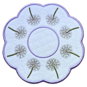 Close up of an embroidered dandelion seed head candle mat with a scalloped purple border, circular centre and delicate dandelion clocks stitched around the edge.