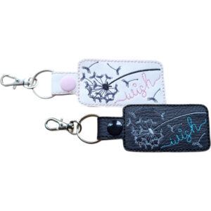 Two embroidered dandelion wish keyrings in white and black faux leather, with silver clips and matching snap fasteners.