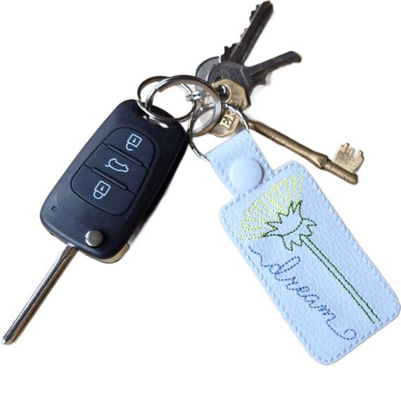 White embroidered dandelion dream keyring attached to a set of keys with car key, showing its size and everyday use.