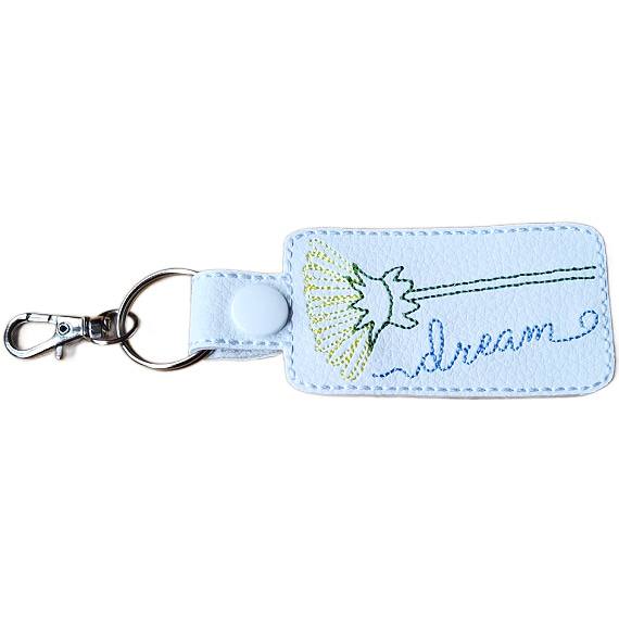 White faux leather dandelion keyring with embroidered flower design, stitched 'dream' wording, white snap fastener and silver keyring clip.