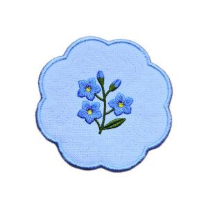 Embroidered forget me not candle coaster with a blue floral sprig in the centre, soft textured background and blue scalloped border.