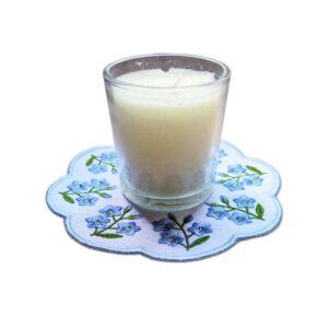 Embroidered forget me not candle mat with delicate blue flowers and green leaves arranged around the edge, shown with a glass candle in the centre.