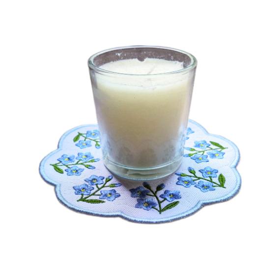 Embroidered forget me not candle mat with delicate blue flowers and green leaves arranged around the edge, shown with a glass candle in the centre.