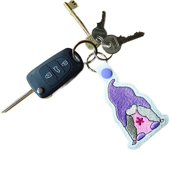 Embroidered girl gnome keyring attached to a set of keys, featuring a purple hat, pink dress, grey hair and sturdy metal hardware.