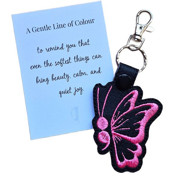 Black faux leather butterfly keyring with pink embroidered wing detail, shown with a matching keepsake card about beauty, calm and quiet joy.