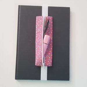 Handmade A4 planner zipper pouch in pink ditsy flower print cotton fabric, shown fitted around a black A4 notebook with a white zip and white elastic strap, holding pens and stationery items.