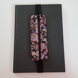 Handmade A4 planner zipper pouch in pink and purple floral print cotton fabric, shown fitted around a black A4 notebook with a black zip and black elastic strap, holding pens and stationery items.