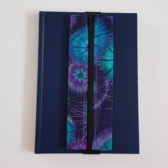 Handmade A5 planner zipper pouch in blue, turquoise and purple tie dye print cotton fabric, shown fitted around a navy A5 notebook with a black zip and black elastic strap, holding pens and stationery items.