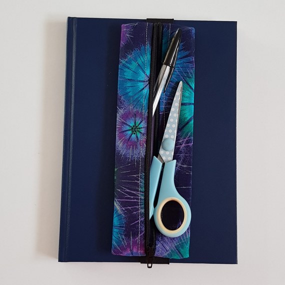 Handmade A5 planner zipper pouch in blue, turquoise and purple tie dye print cotton fabric, shown fitted around a navy A5 notebook with a black zip and black elastic strap, holding pens and stationery items.