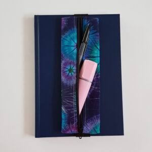 Handmade A5 planner zipper pouch in blue, turquoise and purple tie dye print cotton fabric, shown fitted around a navy A5 notebook with a black zip and black elastic strap, holding pens and stationery items.