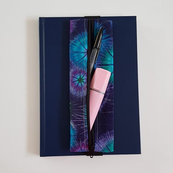 Handmade A5 planner zipper pouch in blue, turquoise and purple tie dye print cotton fabric, shown fitted around a navy A5 notebook with a black zip and black elastic strap, holding pens and stationery items.