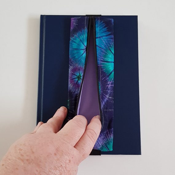 Handmade A5 planner zipper pouch in blue, turquoise and purple tie dye print cotton fabric, shown fitted around a navy A5 notebook with a black zip and black elastic strap, holding pens and stationery items.