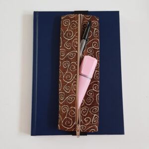 Handmade A5 planner zipper pouch in brown cotton fabric with a cream swirl print, shown fitted around a navy A5 notebook with a matching zip and black elastic strap, holding pens and stationery items.