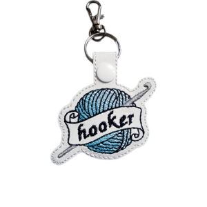 White faux leather keyring embroidered with a teal ball of yarn, silver crochet hook and banner reading 'Hooker', finished with a metal clasp.