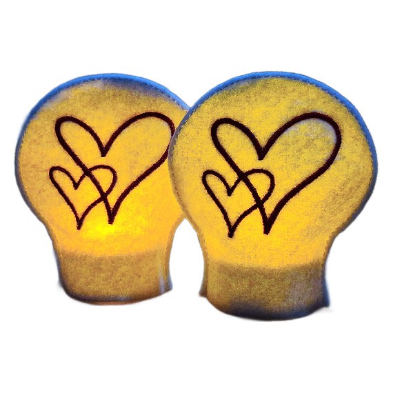 Two lit heart tealight covers glowing warmly over LED tealights, each embroidered with flowing red heart designs on white felt.