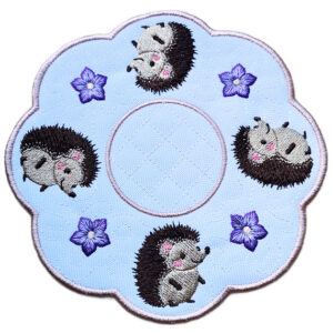 Embroidered hedgehog candle mat close up showing four cute hedgehogs, stitched flower details, scalloped edging and a central circle for a candle or display item.