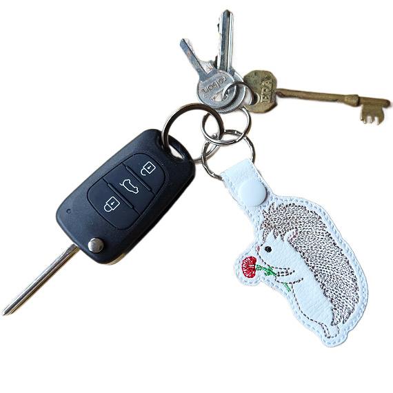 Embroidered hedgehog keyring attached to a set of keys, featuring a cute hedgehog holding a red flower.