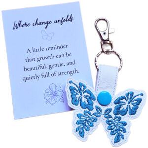 White faux leather hibiscus butterfly keyring with blue floral embroidered wings, shown with a matching keepsake card about growth and change.