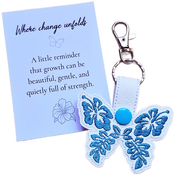 White faux leather hibiscus butterfly keyring with blue floral embroidered wings, shown with a matching keepsake card about growth and change.