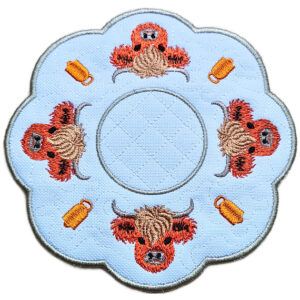 Close up of an embroidered Highland cow candle mat showing four stitched Highland cows, small mug details, scalloped edging and a central circle for a candle or display item.