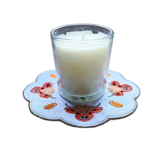 Embroidered Highland cow candle mat with sweet stitched Highland cows, rustic decorative details, scalloped edging and a glass jar candle displayed in the centre.
