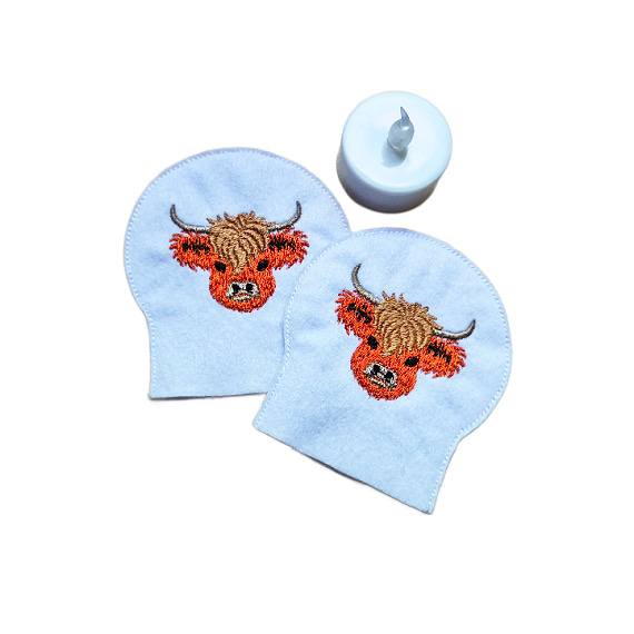Set of two embroidered Highland cow tealight covers made from soft felt, shown with an LED tealight for a cosy farmhouse display.