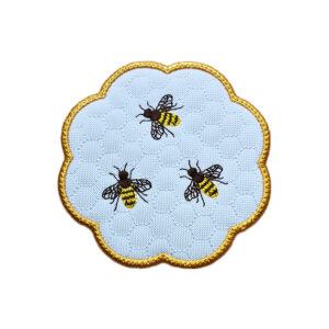 Embroidered honey bee candle coaster on white fabric, with a golden scalloped border, stitched honeycomb background and three detailed bees.