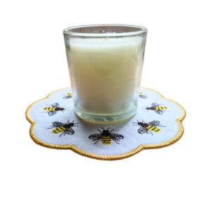 Embroidered honey bee candle mat with a scalloped golden border, shown underneath a glass candle jar with stitched bees around the edge.