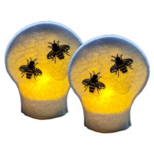 Lit embroidered honey bee tealight covers on white felt, glowing warmly with stitched bees and honeycomb detail over battery operated LED tealights.