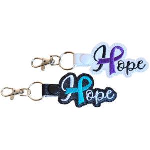 Embroidered Hope ribbon keyrings shown in black and white faux leather, featuring bold stitched Hope wording with purple and teal awareness ribbon details.