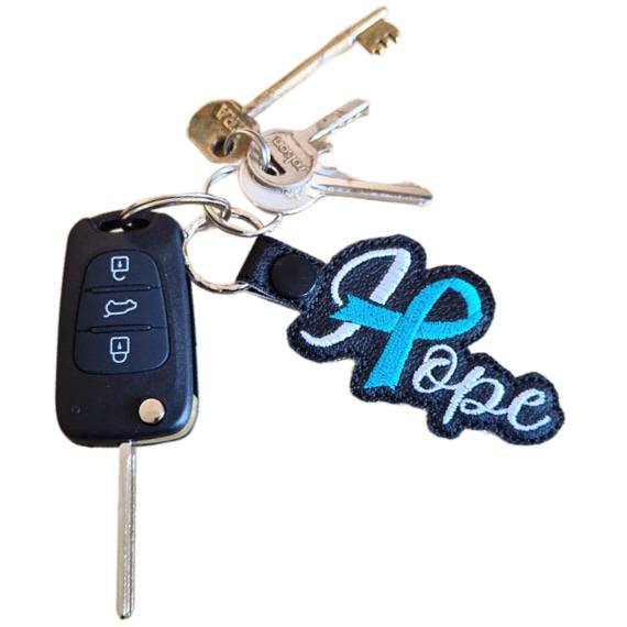 Embroidered Hope ribbon keyring attached to a set of keys, showing the black faux leather design with white stitched wording and a teal awareness ribbon detail.