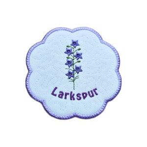 Embroidered July birth flower candle coaster with a purple larkspur stem, stitched larkspur text, background detail and a scalloped purple border.