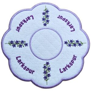 Close up of an embroidered July birth flower candle mat with purple larkspur stems, stitched larkspur text, background detail and a scalloped purple border.