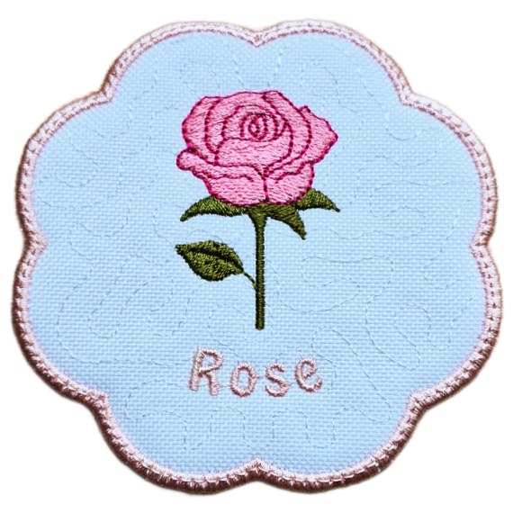 Close up of an embroidered June birth flower candle coaster with a pink rose, green leaves, stitched rose text and a softly scalloped pink border.
