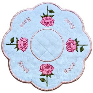 Close up of an embroidered June birth flower candle mat with pink roses, green leaves, stitched rose text and a scalloped pink border.