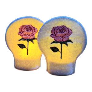 Lit embroidered June birth flower tealight covers on white felt, glowing warmly with pink rose designs over battery operated LED tealights.