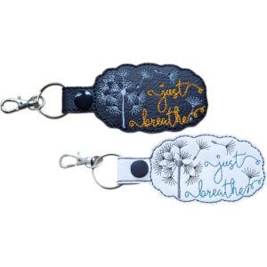 Two embroidered 'just breathe' dandelion keyrings in black and white faux leather, with silver clips and matching snap fasteners.